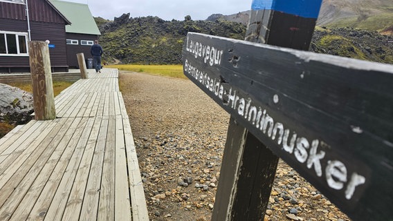 Ein Holzpfad entlang von Holzhütten, daneben ein Holzschild auf isländisch mit der Aufschrift Lavavegur (Lavaweg) © NDR Foto: Merle Schaack und Michael Richmann
