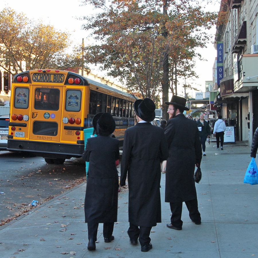 Drei in schwarz gekleidete Männer gehen in den USA auf einem Bürgersteig, daneben ist ein gelber Bus © NDR Foto: Jörn Pissowotzki