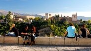 Touristen sitzen auf einer Steinmauer, dahinter sieht man das Gebäude La Alhambra in der Stadt Granada © Instituto de Turismo de España Foto: Juan Serrano