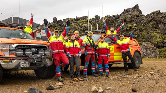 Mehrere jüngere Menschen in auffälligen Rettungsteam-Outfits in Neonfarben stehen winkend vor Rettungsfahrzeugen - sie sind Freiwillige der Rettungswache in Island © NDR Foto: Merle Schaack und Michael Richmann