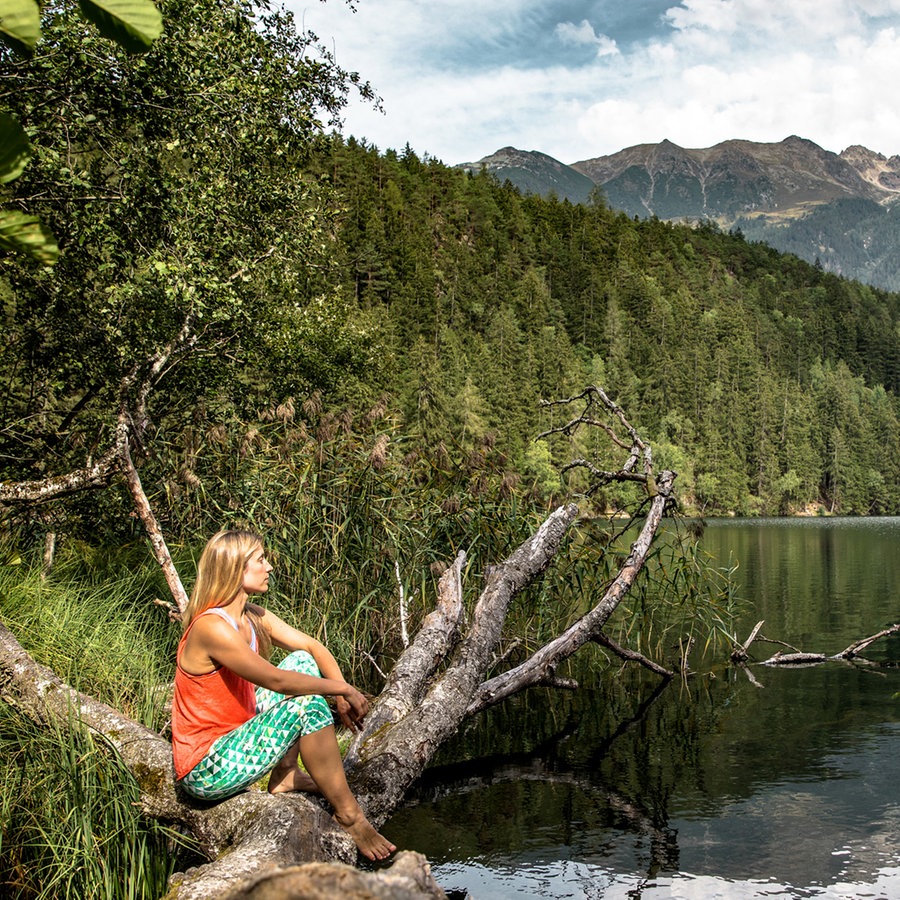 Eine Frau sitzt in lockerer Kleidung auf einem Baumstamm und schaut auf den Pigurber See im Naturpark Öztal © Oesterreich Werbung / The Creating Click.com 