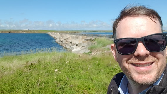 Ein Mann mit Sonnenbrille vor einem Damm auf einer Orkney-Insel © NDR Foto: Thomas H.A. Becker
