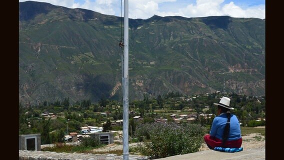 Eine Person mit Hut und blauem Oberteil sitzt auf einem Weg und schaut auf die Landschaft der Anden. © NDR Foto: Anne Passow