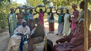 Männer sitzen auf Plastikstühlen, dahinter stehen Frauen mit Kopftüchern (Foto aus dem Senegal) © NDR Foto: Angelika Henkel