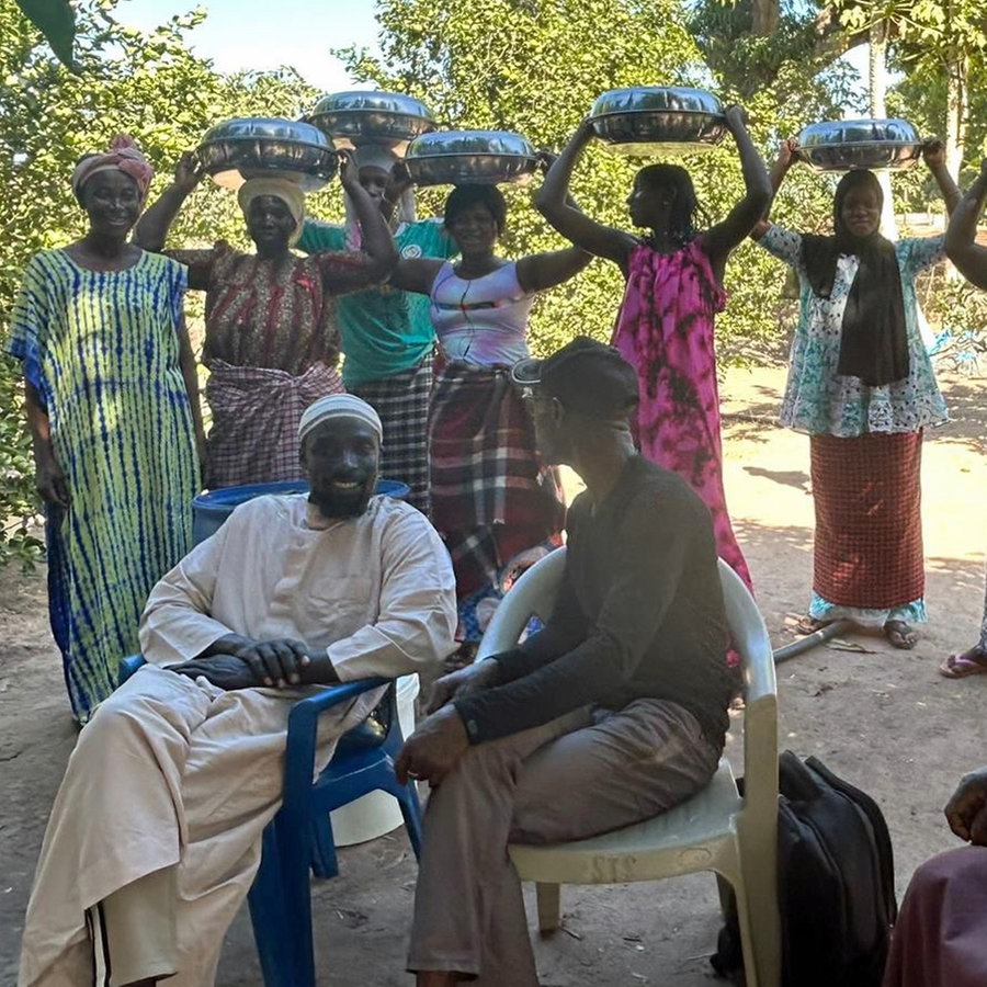 Männer sitzen auf Plastikstühlen, dahinter stehen Frauen mit Kopftüchern (Foto aus dem Senegal) © NDR Foto: Angelika Henkel
