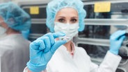 Eine Wissenschaftlerin im Laborkittel mit Mundschutz, Gummihandschuhen und Haarnetz hält ein Glasgefäß hoch. © Colourbox Foto: Arne Trautmann