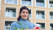 Eine junge Frau steht lächelnd mit einem Umzugskarton in den Händen vor einem Studentenwohnheim. © Colourbox Foto: #221789