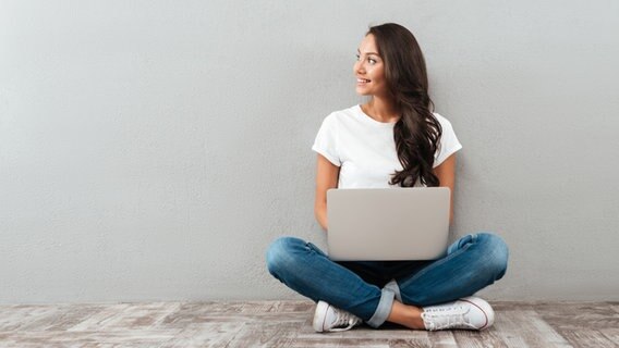 Eine Frau sitzt mit einem Laptop auf dem Schoß auf dem Boden und schaut lächelnd zur Seite. © Colourbox Foto: Pavel Vladychenko