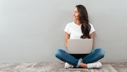 Eine Frau sitzt mit einem Laptop auf dem Schoß auf dem Boden und schaut lächelnd zur Seite. © Colourbox Foto: Pavel Vladychenko