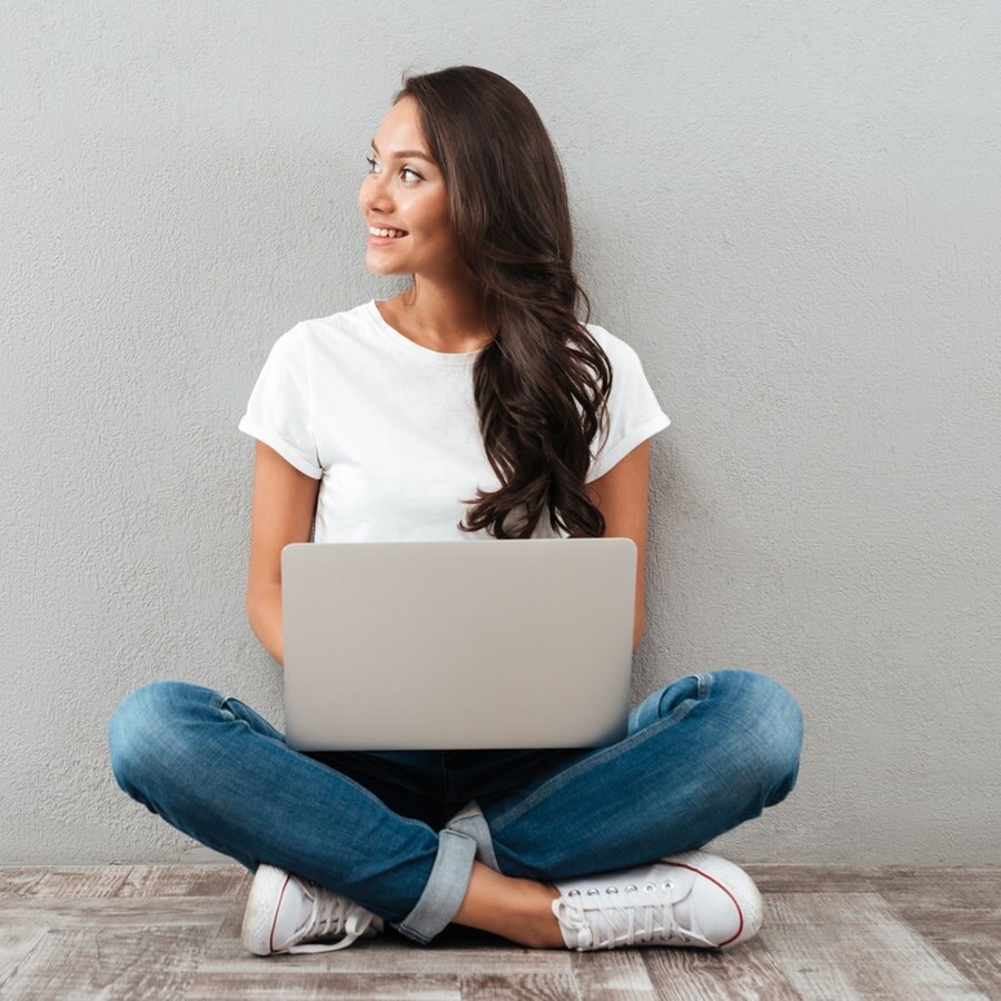 Eine Frau sitzt mit einem Laptop auf dem Schoß auf dem Boden und schaut lächelnd zur Seite. © Colourbox Foto: Pavel Vladychenko