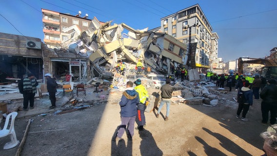 Mitglieder eines Rettungseinsatzteams stehen in den und vor den Trümmern eines bei einem Erdbeben in der Türkei zerstörten Gebäudes und suchen nach Überlebenden. © BRH-Rettungshundestaffel Hamburg und Harburg e.V. Foto: Grinnus