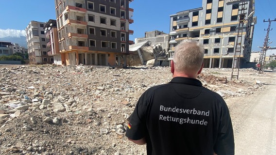 Holger Grinnus vom Bundesverband Rettungshunde steht vor mehreren mehrstöckigen Gebäuden, die beim Erdbeben in der Türkei beschädigt wurden, aber stehen geblieben sind. © BRH-Rettungshundestaffel Hamburg und Harburg e.V. Foto: Grinnus