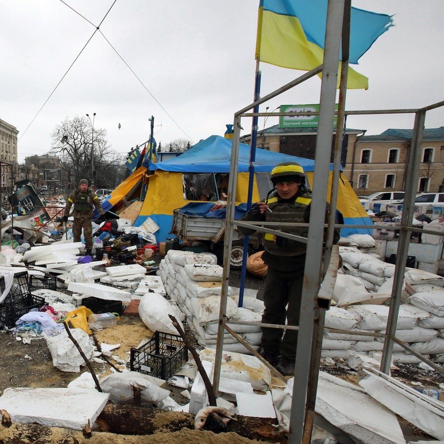 Nach einem Raketeneinschlag vor dem Gebäude der regionalen Verwaltung in Charkiw auf dem Svobody-Platz (Freiheitsplatz) halten sich die Menschen am Zelt der Freiwilligen "Alle für den Sieg" auf, links ist auf einem Plakat das Bild eines Angehörigen des ukrainischen Militärs mit Gesichtsmaske zu sehen. © dpa-Bildfunk 