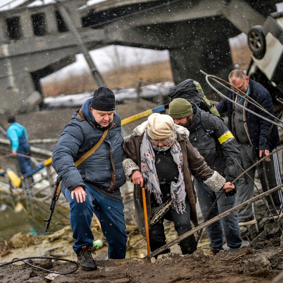 Örtliche Milizionäre helfen einer alten Frau über eine von Artillerie zerstörte Brücke am Stadtrand von Kiew. © picture alliance/dpa/AP | Emilio Morenatti 