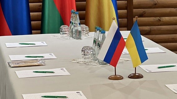 Zwei Fähnchen in den russischen und den ukrainischen Farben stehen auf einem für Friedensverhandlungen vorbereiteten Tisch. © picture alliance / AA | FOREIGN MINISTRY OF BELARUS 
