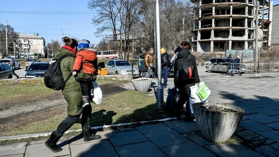 Mehrere Menschen gehen in der ukrainischen Stadt Saporischschja mit Taschen und Tüten in der Hand zu vor einem Haus geparkten Autos. © dpa picture alliance/AA 