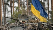 Die ukrainische Flagge ist an einem Radschützenpanzer angebracht, aus dessen Luke ein ukrainischer Soldat guckt. © dpa picture alliance/AP Foto: Vadim Ghirda
