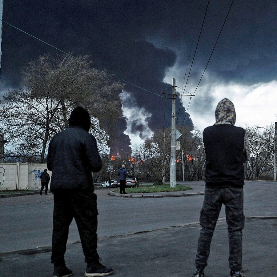 Schwarzer Rauch ist am Himmel über Odessa zu sehen, im Vordergrund stehen vier junge Männer mit dem Rücken zur Kamera und schauen in die Ferne. © imago 