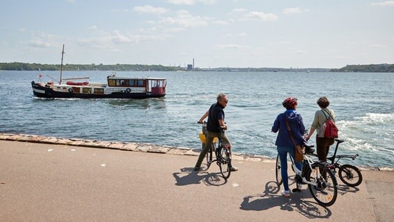 Radfahrer stehen am Tiessenkai in Kiel-Holtenau während ein Schiff vorbeifährt. © dpa-Bildfunk Foto: Georg Wendt/dpa