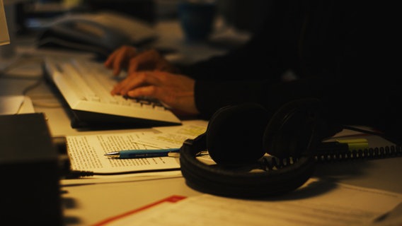 Eine Person sitzt an einem spärlich beleuchteten Schreibtisch und tippt auf einer Tastatur. © NDR 