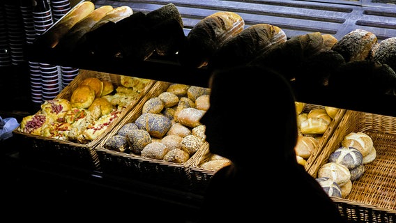 Eine Bäckereifachverkäuferin steht am frühen Morgen in einer sparsam beleuchteten Bäckerei. Im dunklen Verkaufsraum sind nur die Umrisse der Frau vor dem beleuchteten Brötchenregal zu sehen. © dpa Foto: Julian Stratenschulte