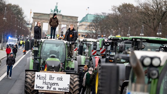 Landwirte stehen mit ihren Traktoren in einer Schlange vor dem Brandenburger Tor in Berlin. Auf einem Plakat an der Front eines Traktors, auf dem auch drei Teilnehmer stehen, steht die Aufschrift "Müsst ihr erst hungern, bevor ihr es versteht?". © dpa Foto: Fabian Sommer