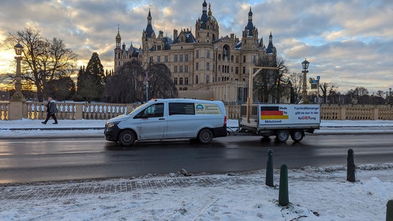 Vor dem Schweriner Schloss steht ein Kleintransporter mit einem Anhänger mit der Aufschrift "Made by Mittelstand" neben einer Deutschlandflagge. Im Hintergrund ein idyllisches Bild: Das Schloss wird von der Sonne, die zwischen einigen Wolken durchdringt, golden angestrahlt. © NDR Foto: Christoph Loose