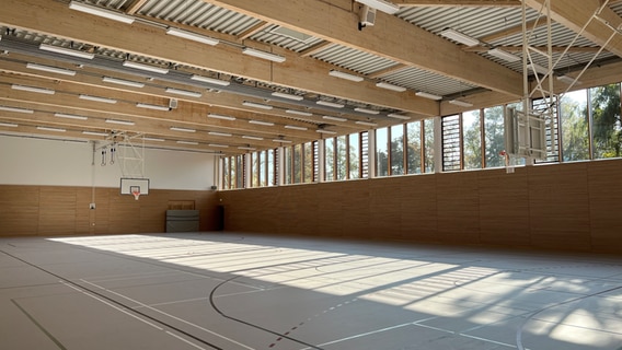 Die nagelneue Sporthalle des Grundschulzentrums Boizenburg. Zwei Basketballkörbe hängen an weißem Gestänge von der Decke. Durch eine große Fensterfront fällt helles Licht in die Halle und auf deren hellgrauen, mit frischen Markierungslinien durchzogenen Boden. Das Dach wird von massiven Holzbalken gestützt, dazwischen hängen parallel zu den Balken weiße, längliche Neonleuchten. Die Hallenwände sind von unten bis zur halben Höhe mit Holz verkleidet, die obere Hälfte ist weiß gestrichen. Die nagelneue Sporthalle des Grundschulzentrums Boizenburg. Zwei Basketballkörbe hängen an weißem Gestänge von der Decke. Durch eine große Fensterfront fällt helles Licht in die Halle und auf deren hellgrauen, mit frischen Markierungslinien durchzogenen Boden. Das Dach wird von massiven Holzbalken gestützt, dazwischen hängen parallel zu den Balken weiße, längliche Neonleuchten. Die Hallenwände sind von unten bis zur halben Höhe mit Holz verkleidet, die obere Hälfte ist weiß gestrichen. © NDR Foto: Lucie Kluth