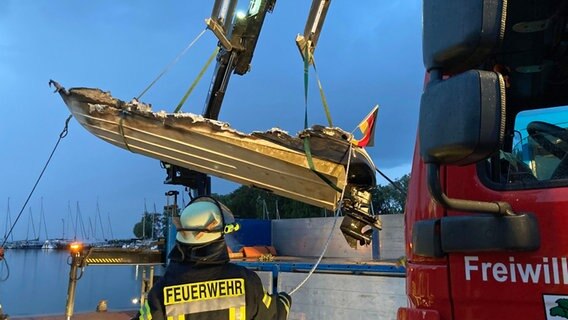 Ein bis zum Rumpf verkohltes Sportboot wird aus dem Wasser gehoben, am Heck weht die Deutschlandfahne, im Hafen von Mönkebude - im Vordergrund ein Feuerwehrmann und ein Feuerwehrauto  Foto: Christopher Niemann