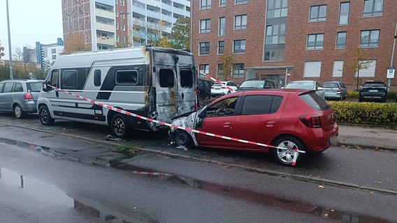 Zwei sichtlich durch einen Brand beschädigte Fahrzeuge - ein Auto und ein Wohnmobil - stehen hintereinander von Absperrband umrahmt am Straßenrand. © NonstopNews Rostock / Stefan Tretropp Foto: Stefan Tretropp