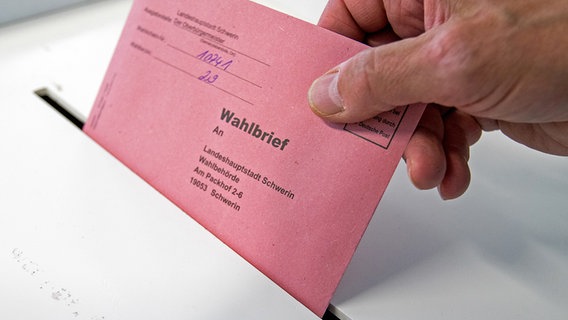 Eine Hand hält einen rosafarbenen Briefumschlag, der an der Unterseite bereits im Schlitz einer Wahlurne steckt. Er ist beschriftet mit "Wahlbrief". © dpa Foto: Jens Büttner