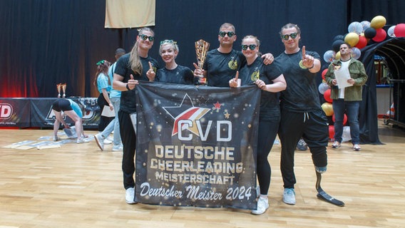 Die Cheerleader des Cheer-Company-Greifswald e.V. tragen Kostüme und herzförmige Sonnenbrillen und lächeln in die Kamera. Sie halten ein Banner mit der Aufschrift "CVD - Deutsche Cheerleading Meisterschaft - Deutscher Meister 2024". © Cheer-Company-Greifswald e.V. Foto: Cheer-Company-Greifswald e.V.