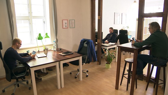 Drei Männer sitzen in einem Büro, jeder an einem Tisch.  