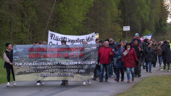 Rund 170 Menschen haben sich zu einem Protestmarsch in der Gemeinde Dabel versammelt. Sie demonstrieren gegen eine geplante Geflüchtetenunterkunft und gehen Banner schwingend über einen asphaltierten Weg im Dorf. © NDR Foto: NDR