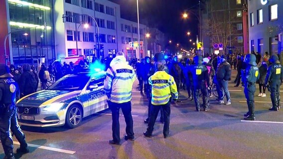 Auf einer Straßenkreuzung in Rostock steht ein Polizeiwagen mit Blaulicht. Im Hintergrund ziehen Demonstranten vorbei, im Vordergrund sind mehrere Polizisten zu sehen. © TeleNewsNetwork Foto: TeleNewsNetwork
