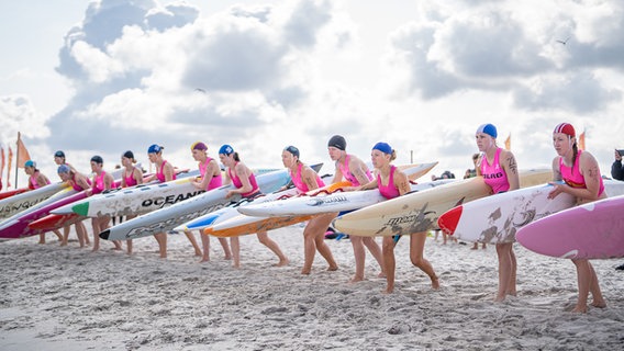 13 Rettungsschwimmerinnen stehen vor sonnig-wolkigem Himmel in einer Reihe am Strand. Sie tragen pinke Badeanzüge mit dem DLRG-Logo und Badekappen. Offensichtlich beginnt gleich ein Rennen - die Frauen befinden sich in lauernder Stellung und haben ihre Surfbretter unter den Arm geklemmt. © DLRG Foto: DLRG