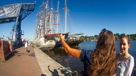 Zwei Frauen machen ein Selfie vor einem Segelschiff im Rostocker Stadthafen zur Hanse Sail 2022. © dpa Foto: Frank Hormann