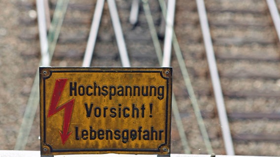 Ein Schild mit einem Blitz und der Aufschrift "Hochspannung. Vorsicht! Lebensgefahr." Im Hintergrund Bahnschienen. © imago/Waldmüller Foto: imago/Waldmüller