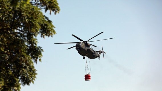 Ein Hubschrauber der Bundeswehr fliegt Wasser in einem angehängten Behälter zu Brandstellen im Gördenitzer Moor. © NonstopNews Rostock Foto: Stefan Tretropp