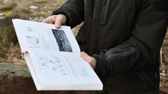 Eine Nahaufnahme: Ein Mann hält ein aufgeschlagenes Buch in den Händen. Zu sehen sind Fotos, zeichnungen und Erläuterungen zu einem Hügelgrab. © NDR Foto: Janet Lindemann