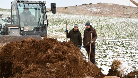 Zwei Männer, René Geyer und Johannes Stiller, stehen mit Schaufeln auf einem leicht verschneiten Feld hinter einem Haufen von Erde. Links daneben wird ein Radlader rangiert. © NDR Foto: Janet Lindemann