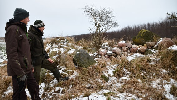 Zwei Männer, René Geyer und Johannes Stiller, stehen auf einem leicht verschneiten Feld neben einem Hügelgrab, ein kleiner Hügel aus Steinen und Erde. Sie unterhalten sich und blicken dabei auf das Grab. © NDR Foto: Janet Lindemann