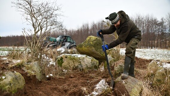Mithilfe eines Spatens stemmt ein Mann einen Stein von einem Hügelgrab. © NDR Foto: Janet Lindemann