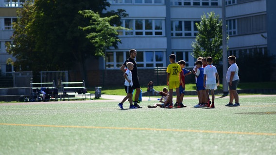Eine Traube von Kindern steht in Sportkleidung um einen Trainer auf einem Kunstrasenplatz. Die Sonne scheint, im Hintergrund ist ein größeres Gebäude zu sehen. © IFC Rostock Foto: IFC Rostock