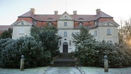 Die Front des 1731 errichteten Barockschlosses in Karlsburg. Ein verschneiter, von zwei Büsten flankierter Kiesweg führt zu einer Treppe und schließlich zur schwarzen, hölzernen Eingangstür des hellen Baus mit Ziegeldach. Die Treppe wird von Nadelbäumen eingerahmt. © dpa Foto: Stefan Sauer