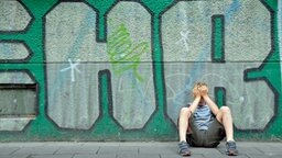 Ein Junge sitzt mit den Händen vor dem Gesicht an einer Häuserwand voller Graffiti. © Chromorange Foto: Walter G. Allgöwer