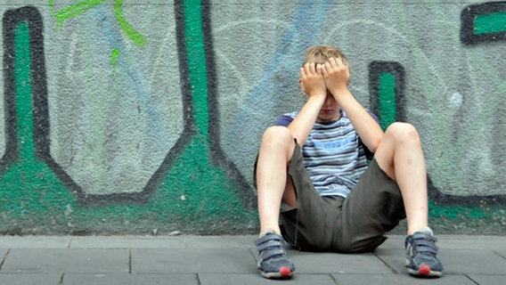 Ein Junge sitzt mit den Händen vor dem Gesicht an einer Häuserwand voller Graffiti. © Chromorange Foto: Walter G. Allgöwer