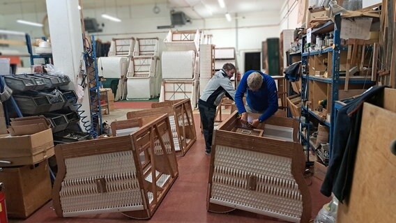 Zwei Mitarbeiter von Korbwerk arbeiten in der Strandkorb-Herstellung, hantieren an je einem Korb in einem größeren Raum voller Körbe in unterschiedlichen Stadien der Herstellung. © NDR Foto: Dörte Rochow