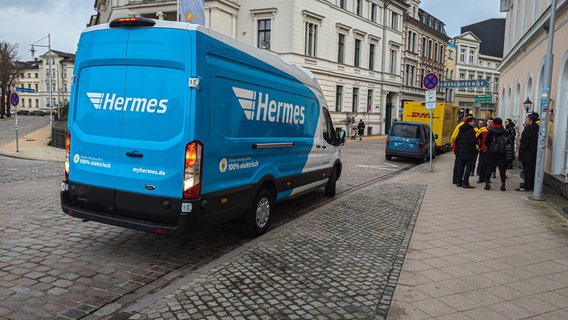 Ein Transporter des Lieferdienstes Hermes mit der bekannten weiß-blauen Farbe steht am Straßenrand in der Schweriner Arsenalstraße. Daneben eine Gruppe von Menschen, im Hintergrund steht ein weiterer Transporter des Lieferdienstes DHL. © NDR Foto: Christoph Loose