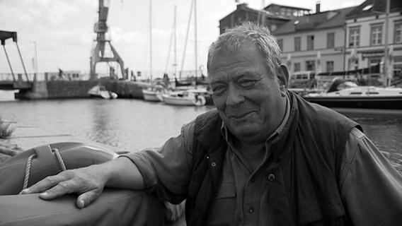 Ein Mann steht an ein Geländer gelehnt. Im Hintergrund ist ein Hafen zu sehen. Es ist der Schauspieler und Autor Lutz Riemann. © NDR Foto: NDR